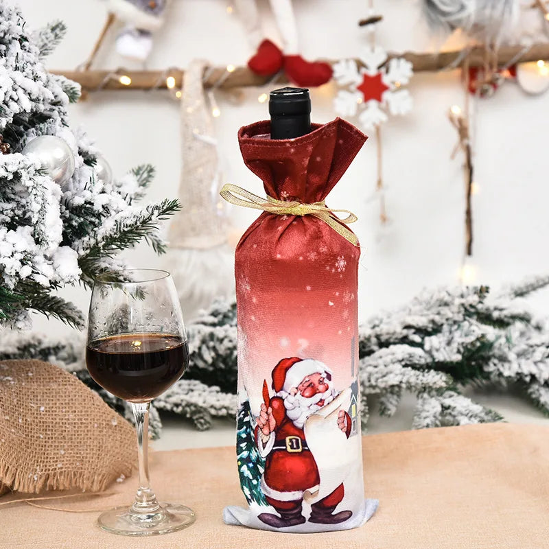 A Christmas-themed wine bottle with a Santa cover next to a glass of red wine, surrounded by festive decor and snow-dusted branches on a beige tablecloth.