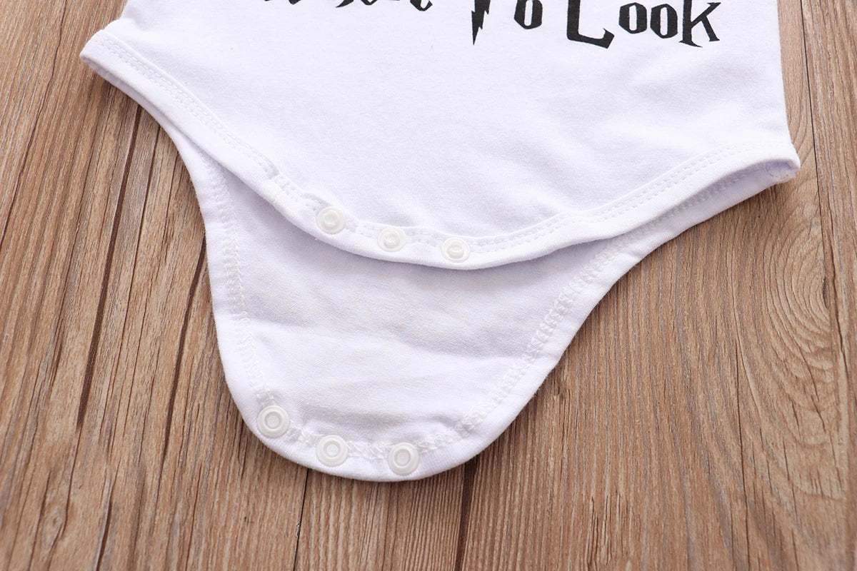 White baby onesie with black text partially visible, featuring three plastic snap buttons at the bottom. It is laid flat on a wooden surface.