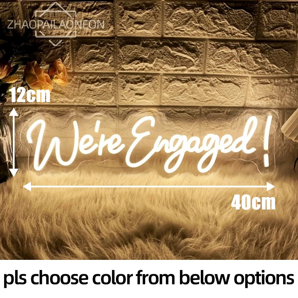 Neon sign reading "We're Engaged!" in warm white light, measuring 12cm by 40cm, set against a brick wall with a fluffy rug below. Flowers are partially visible on the left.