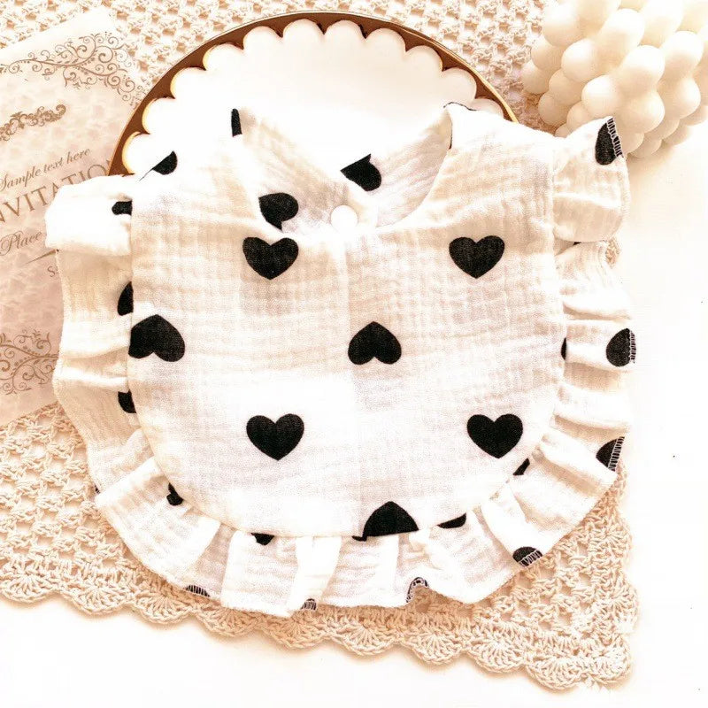White ruffled baby bib with black heart patterns, placed on a decorative plate and crocheted mat. Adjacent is an elegant card and white spherical decorations.