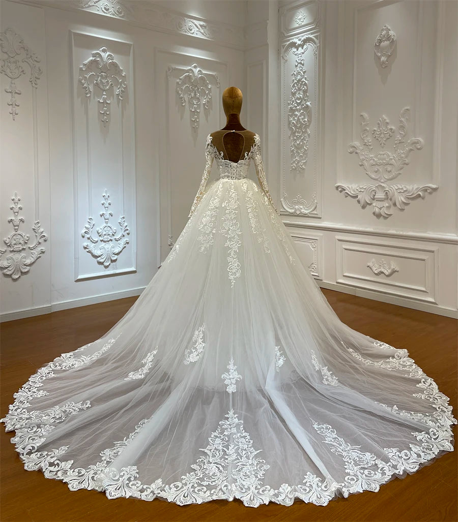 A mannequin displays a long-sleeved, lace wedding gown with an intricate embroidered train, set against an ornate white wall with decorative molding.