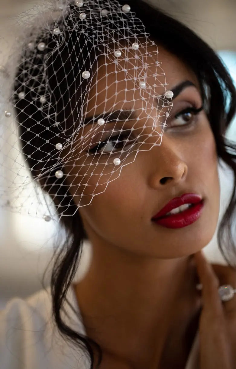Woman with red lipstick wearing a pearl-accented net veil, gazing thoughtfully to the side. Her dark hair is styled back, and she gently touches her neck.