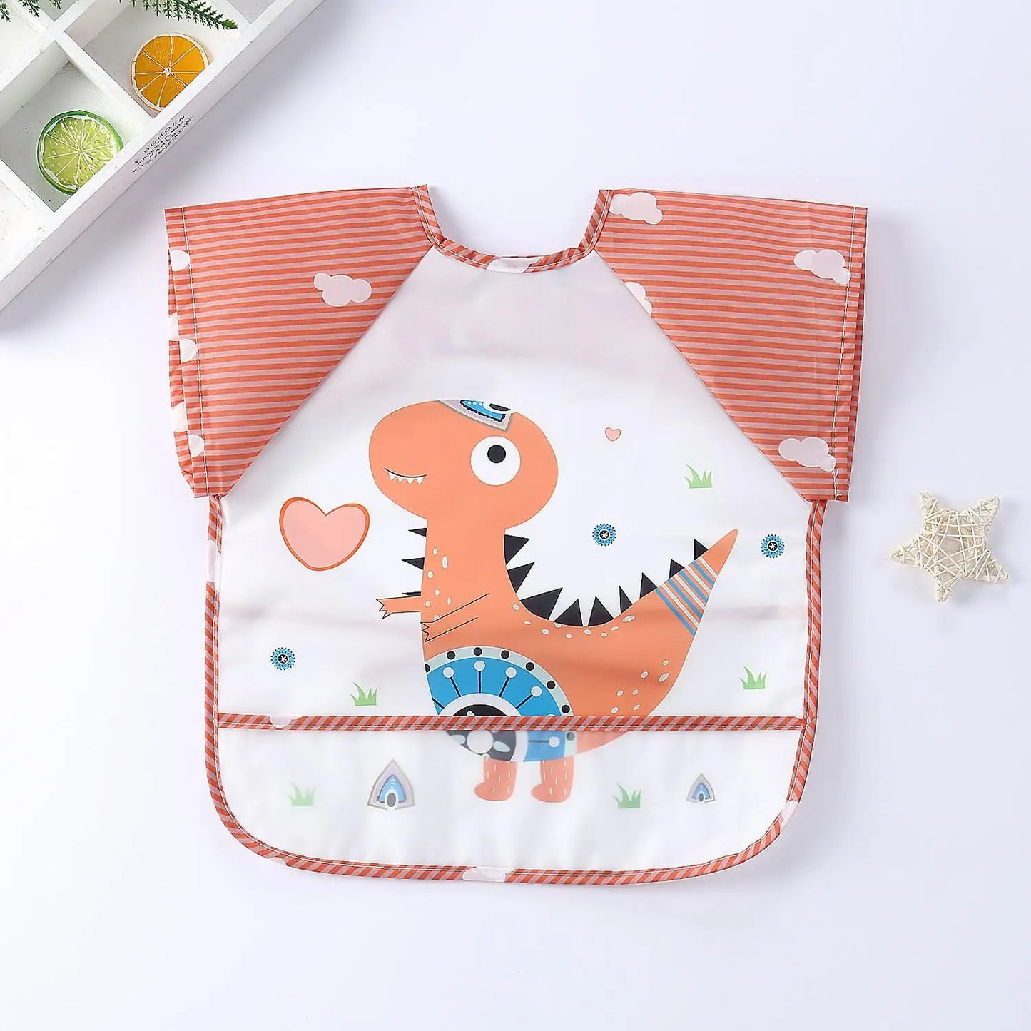Orange-striped bib with a cute cartoon dinosaur, heart, and cloud designs on a white background. A decorative tray with citrus slices is partly visible in the top corner.