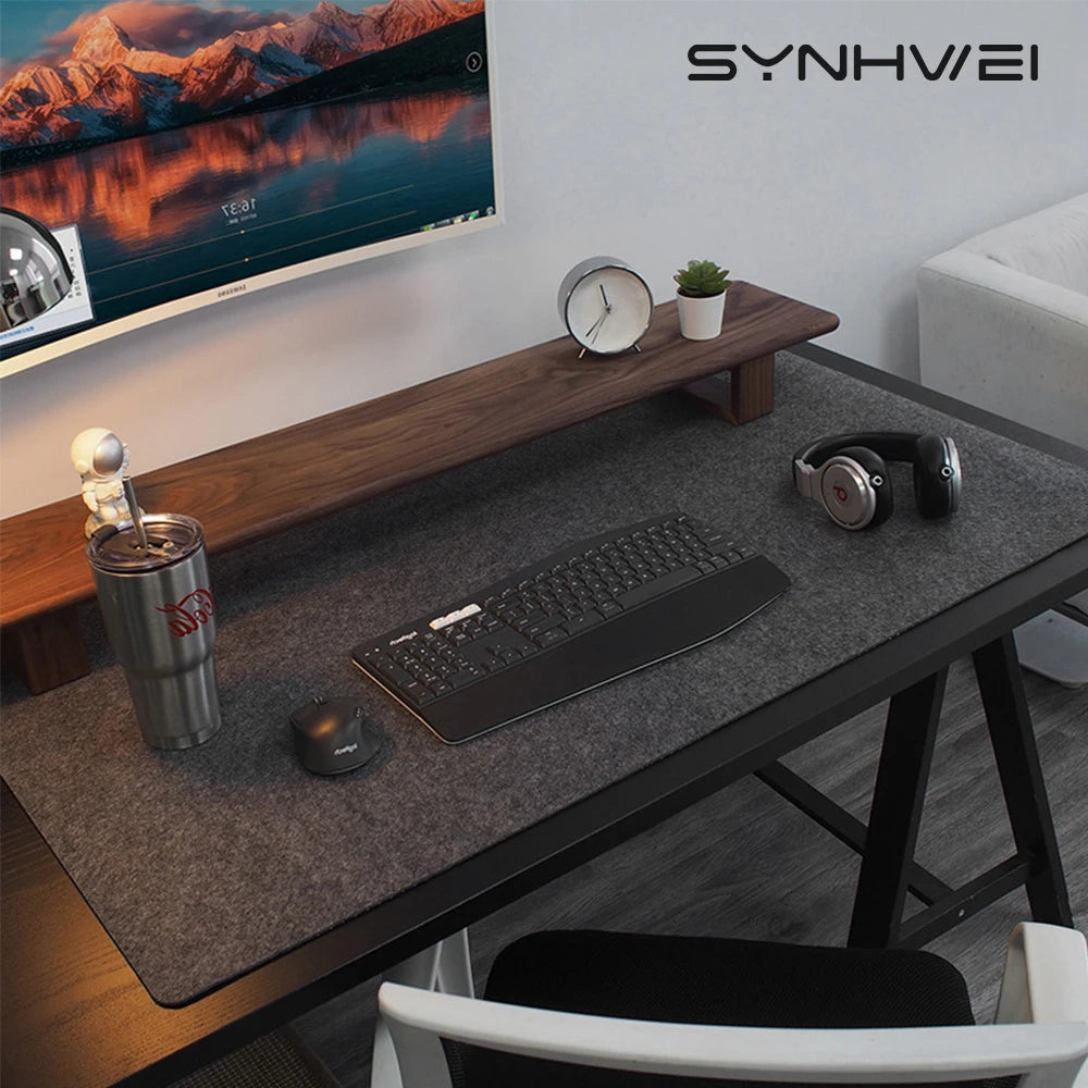 Modern desk setup with a large monitor, wooden stand, keyboard, mouse, headphones, and a tumbler with a small astronaut figurine. A clock and plant sit on the stand.