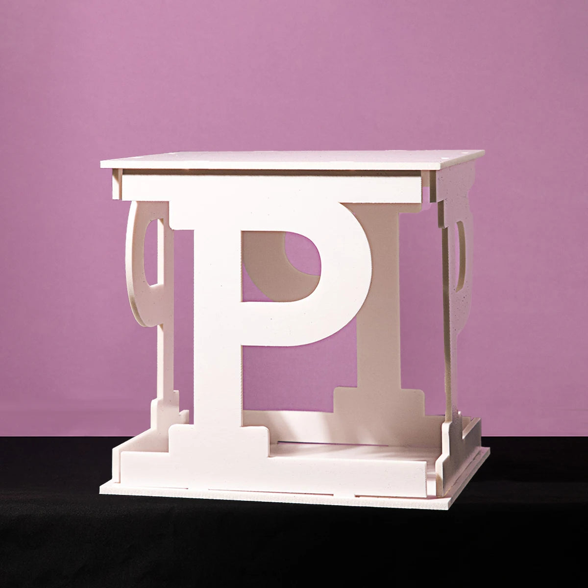 A white, cube-shaped sculpture resembling the letter "P" on multiple sides sits on a black surface against a pink background.