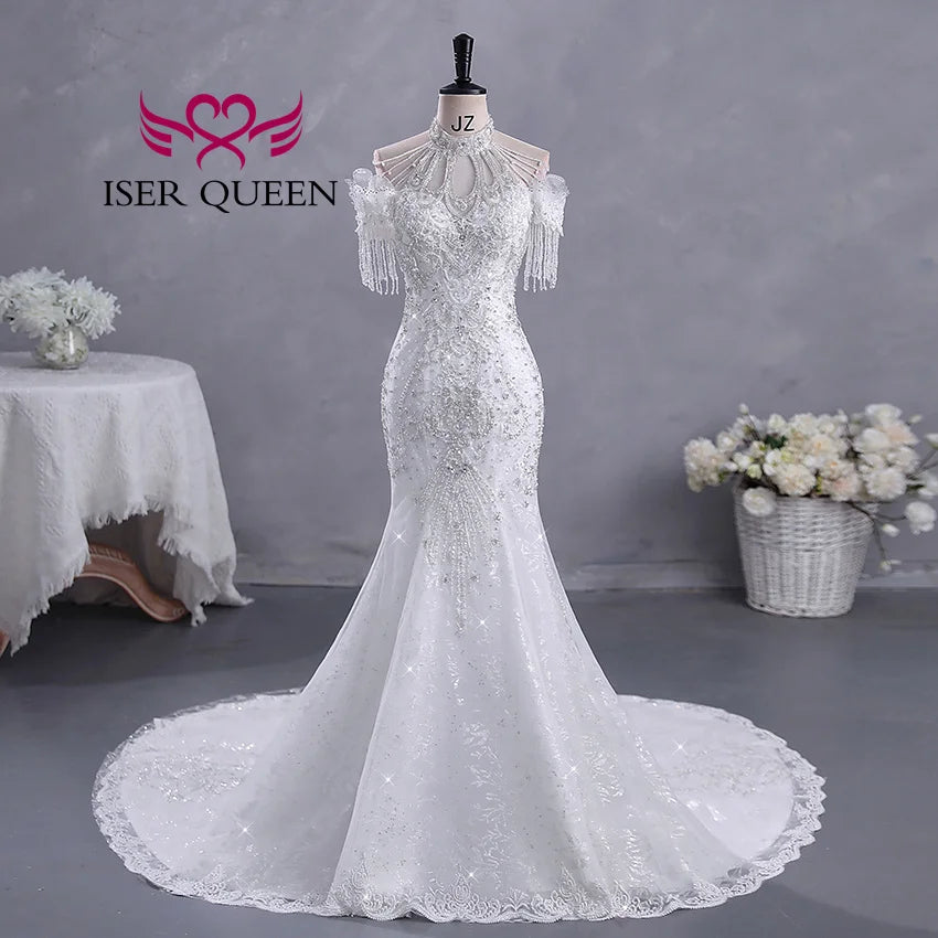 Elegant white wedding dress displayed on a mannequin, featuring intricate lace, beading, and off-shoulder fringe sleeves. Surrounded by a table and flowers in a basket.