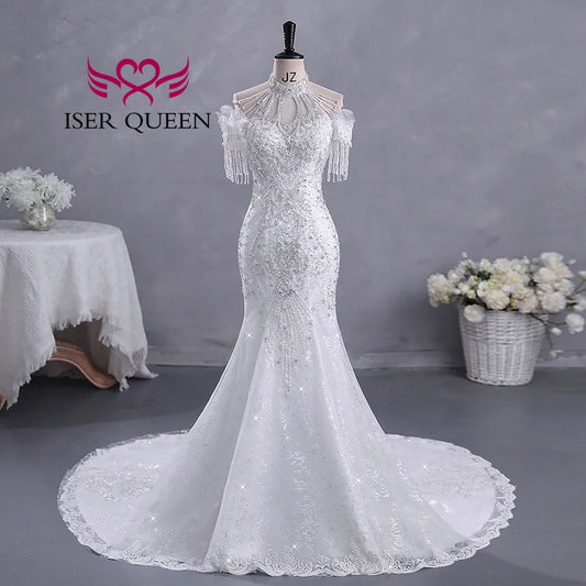 Elegant white wedding dress displayed on a mannequin, featuring intricate lace, beading, and off-shoulder fringe sleeves. Surrounded by a table and flowers in a basket.