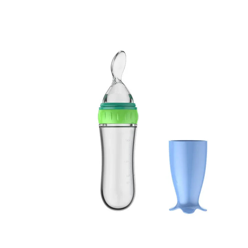 Transparent baby feeding bottle with a green lid and spoon attachment, next to a detachable blue cover on a white background.