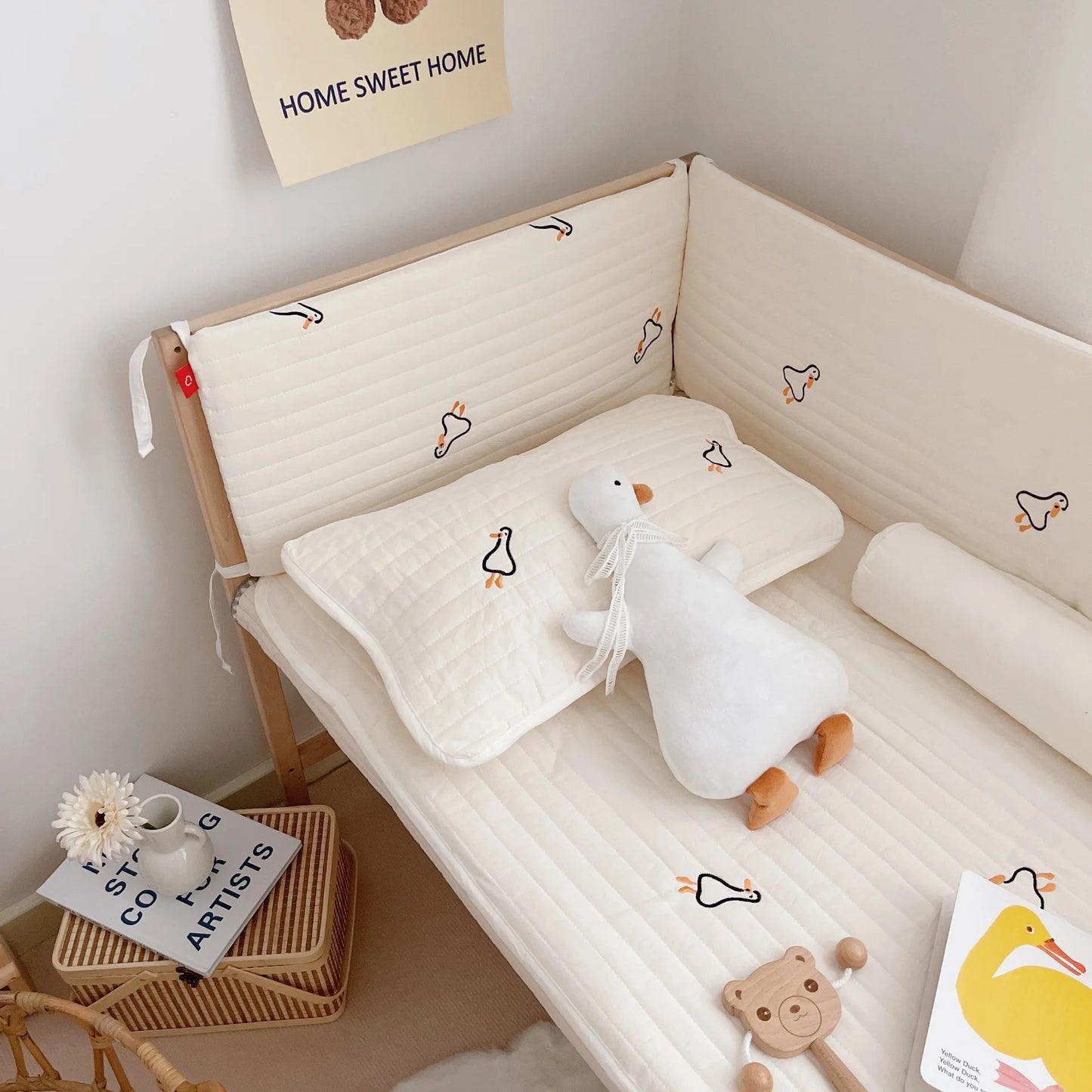 A cozy nursery corner with a beige crib featuring duck-patterned bedding, a plush duck toy, and a small table holding a flower, mug, books, and a "Home Sweet Home" poster on the wall.
