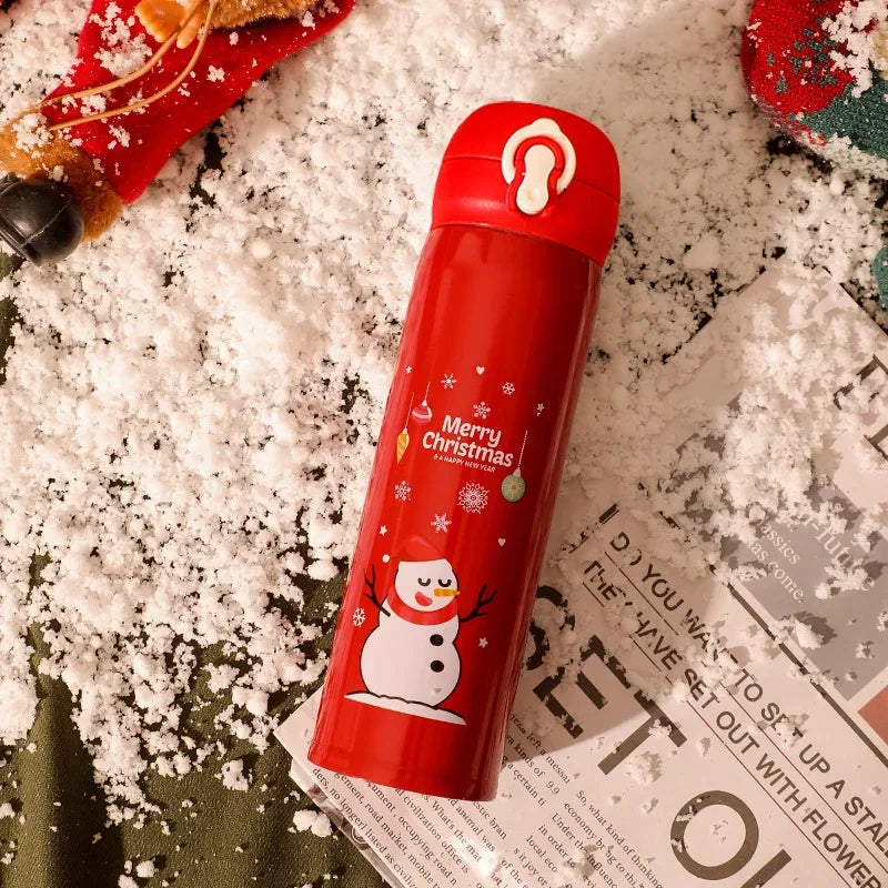 Red thermos with "Merry Christmas" and snowman design lies on fake snow, surrounded by holiday decorations and a magazine.