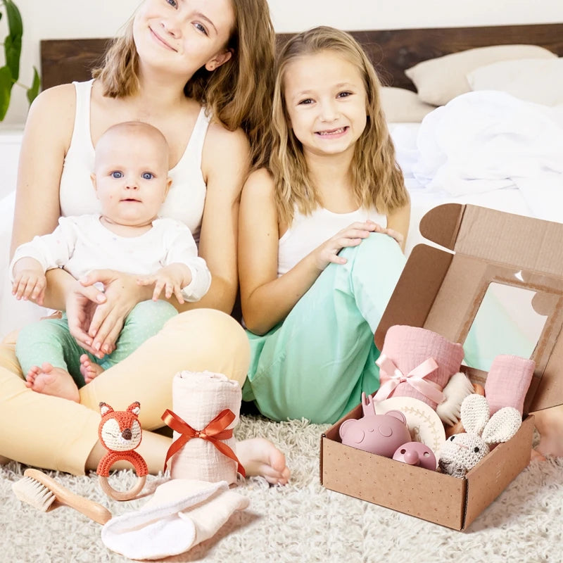 A woman and two children sit on a bed with a gift box nearby, containing plush toys and baby items. The woman holds a baby wearing green pants, while the older child sits beside her.