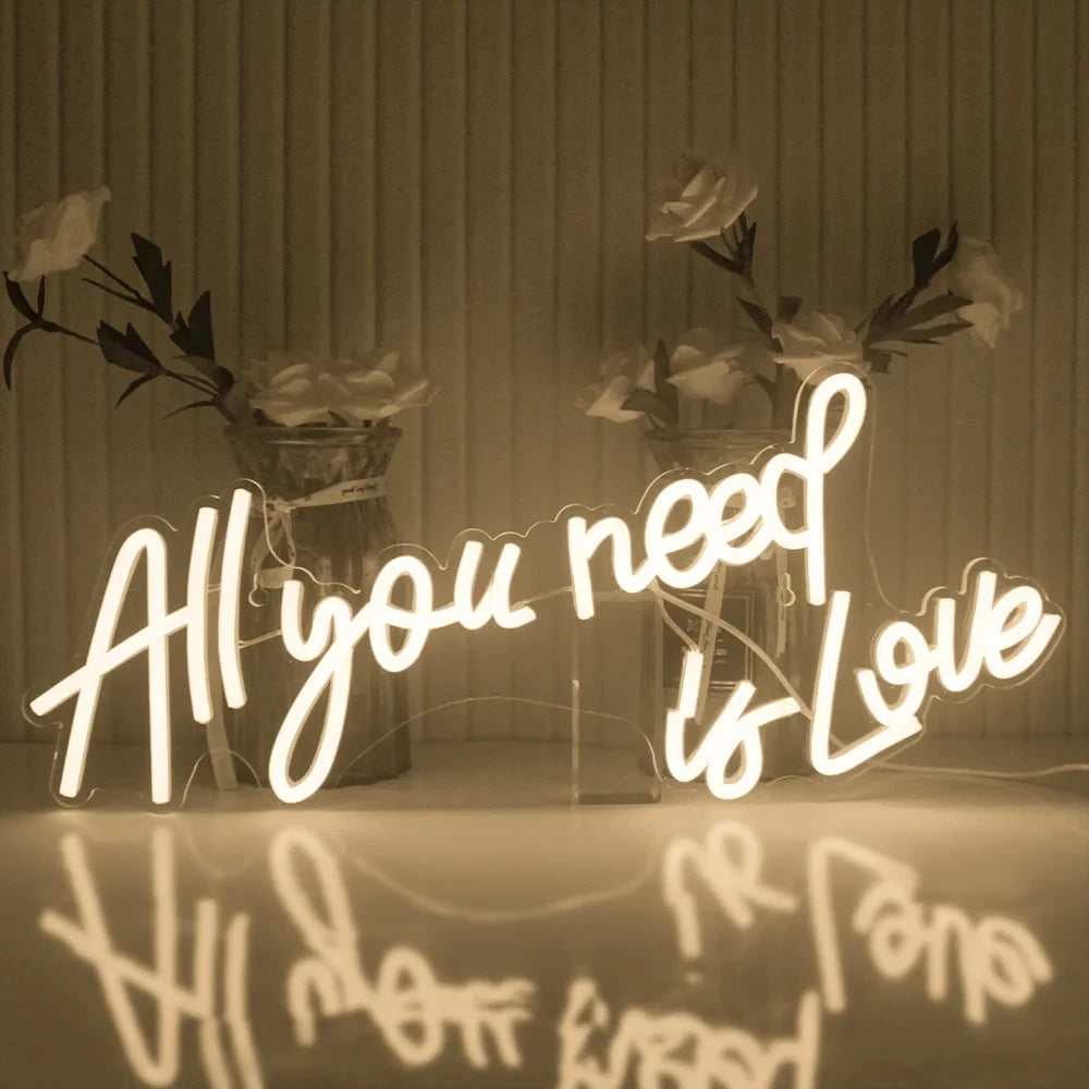 Neon sign reading "All you need is Love" glows warmly against a backdrop of vases with flowers, with its reflection visible on a glossy surface below.