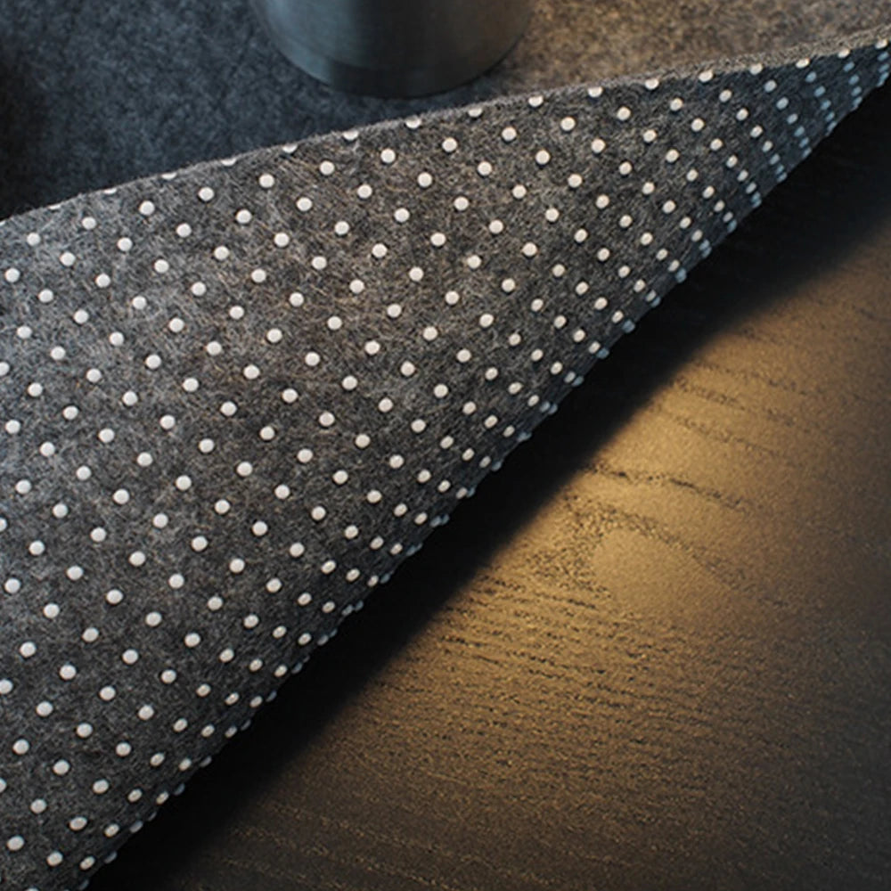 Close-up of a gray anti-slip mat with white dots on a dark wooden surface. The corner of the mat is slightly lifted, revealing its texture and pattern.