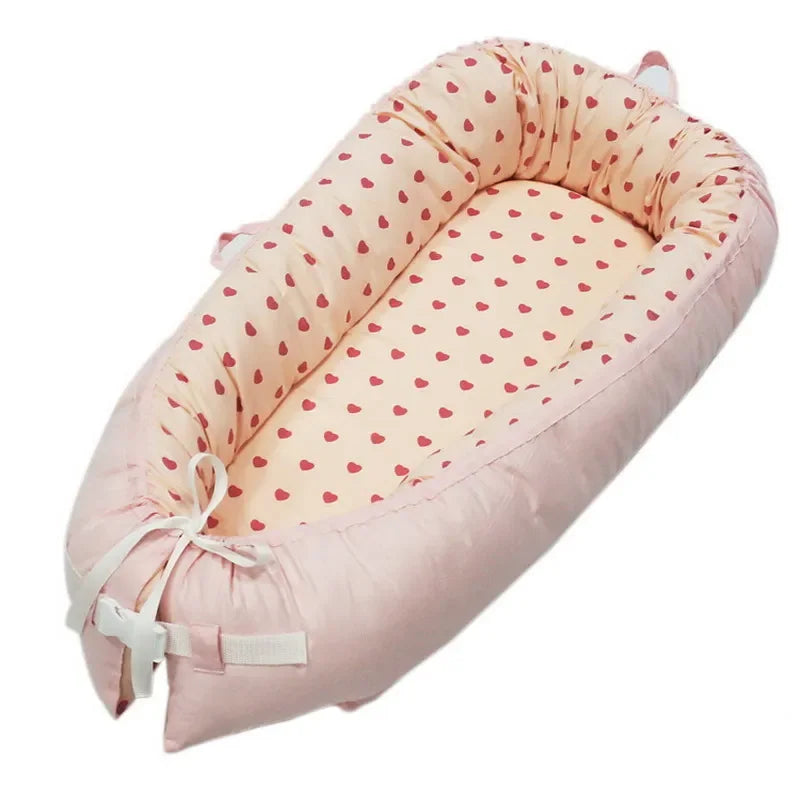 Pink baby nest with a cream interior, adorned with small red hearts. Soft, padded sides and tie closures provide a cozy, secure space.