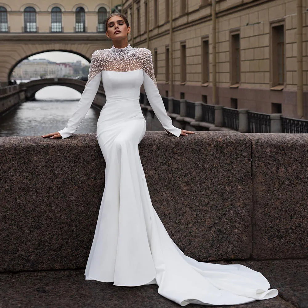 Woman in sleek white gown with pearl-encrusted neckline, sitting on a stone ledge beside a canal. Background shows a bridge and classical architecture.