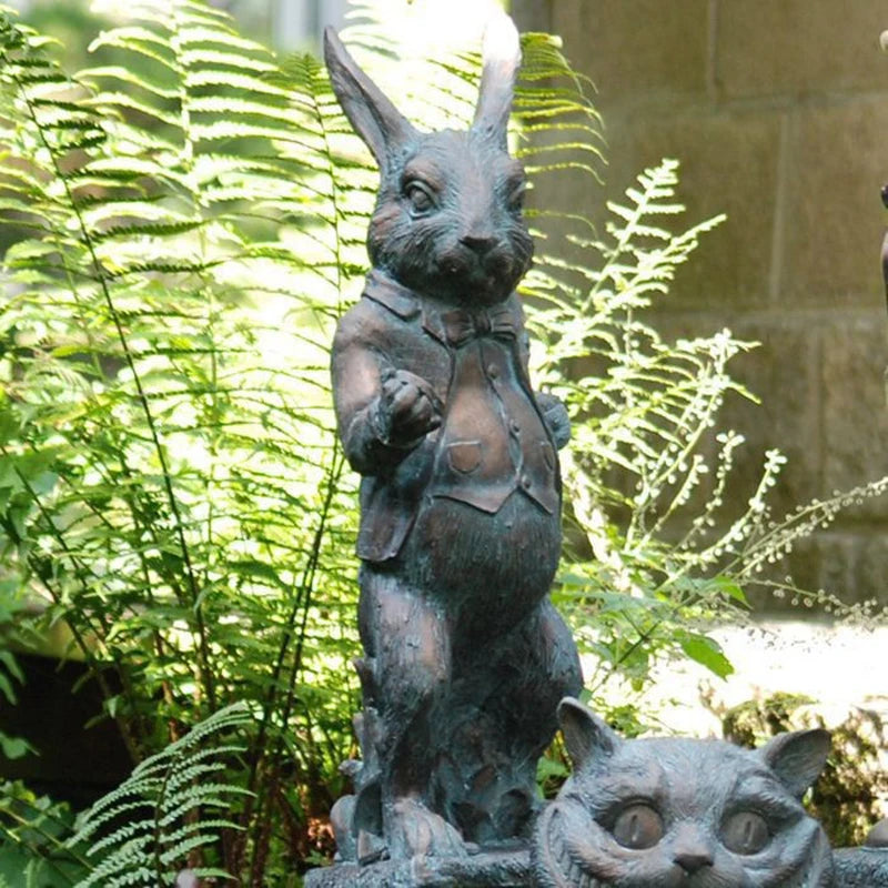 Bronze statue of a rabbit in a suit, standing above a cat head, surrounded by lush green ferns with sunlight filtering through.