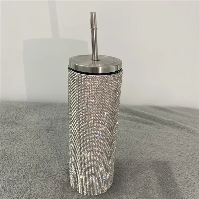 A tall, cylindrical tumbler covered in sparkling rhinestones with a stainless steel lid and straw, placed on a soft gray surface.