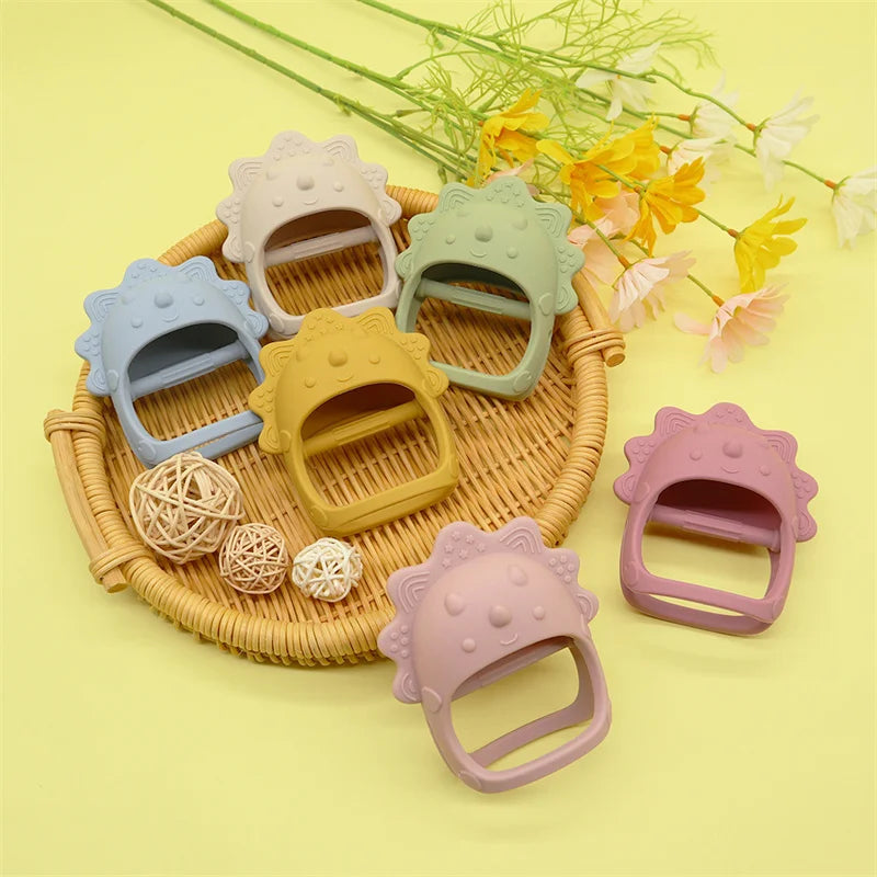 Colorful silicone baby teethers shaped like cute dinosaurs, arranged on a wicker tray with decorative balls and surrounded by flowers on a yellow background.