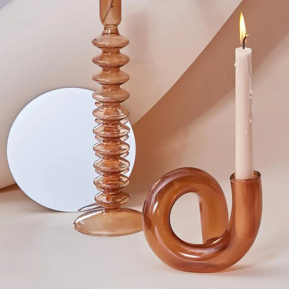 Abstract candle holders and a mirror on a beige surface. One holder spirals with a tall candle lit, casting a shadow. The second is a textured, vertical design nearby.
