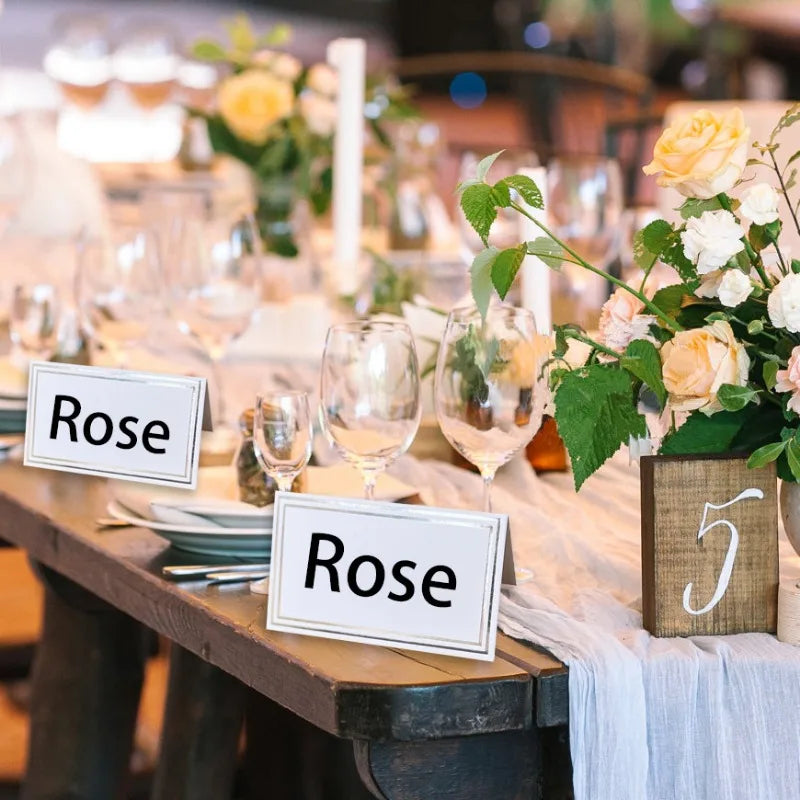 Elegant table setting with floral centerpieces and glassware, featuring name cards labeled "Rose" and a wooden sign with the number five, on a rustic wooden table.