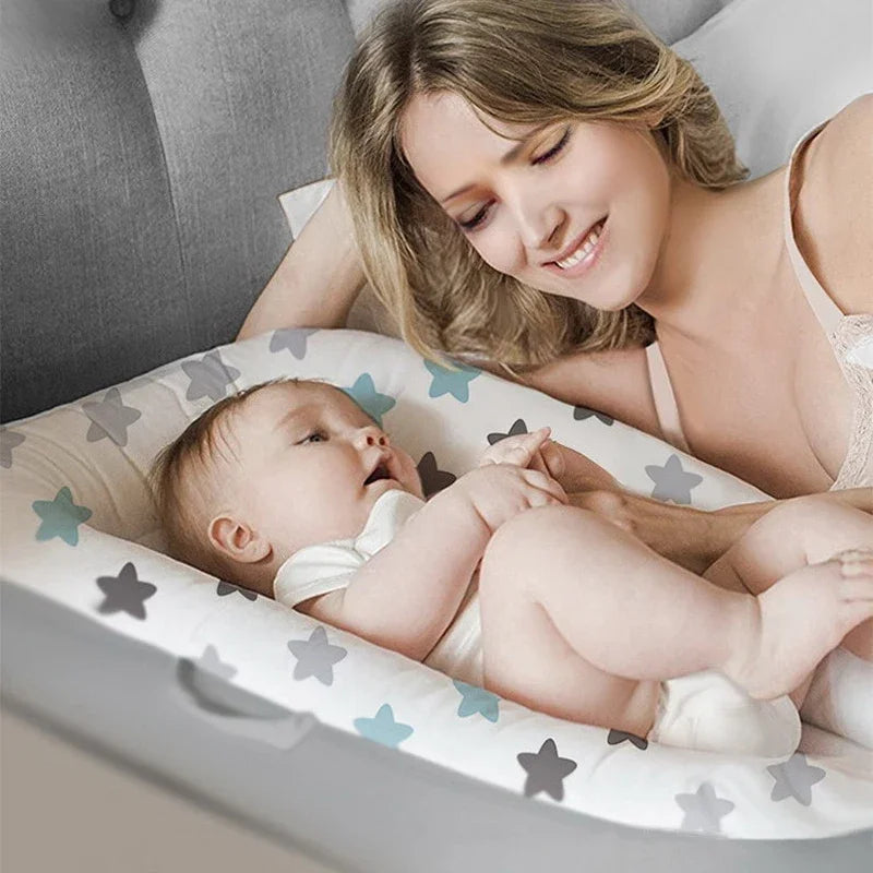 A smiling woman lies in bed, gazing lovingly at a baby in a star-patterned baby nest. The baby, wearing a white onesie, looks up at her, surrounded by soft gray and blue stars.