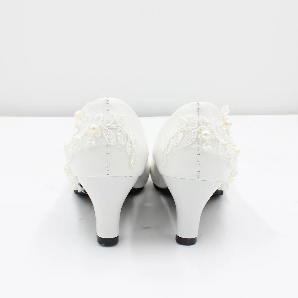 White high-heeled shoes adorned with delicate lace embroidery and small pearls, viewed from the back against a plain white background.