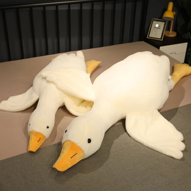 Two large plush white ducks with yellow beaks and feet are laying on a bed. A bedside table with a lamp and picture frame is visible in the background.