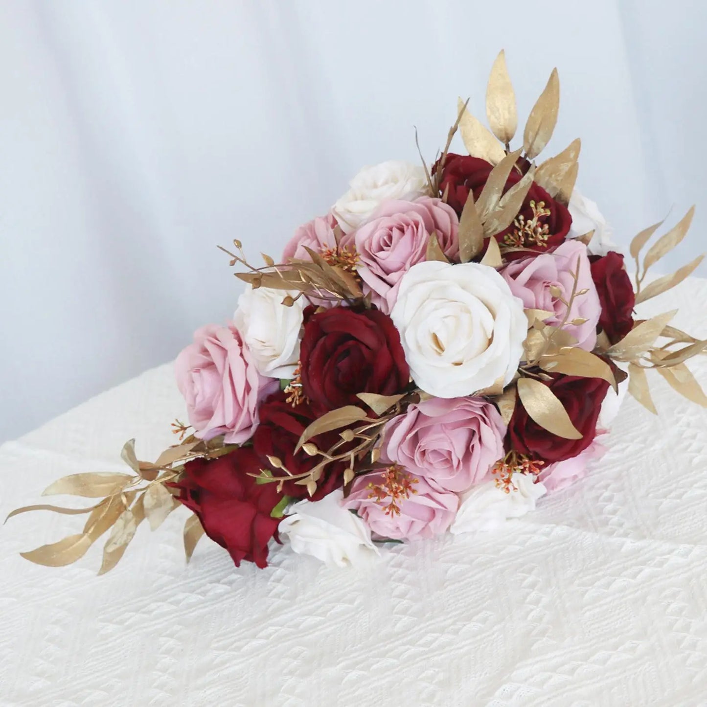 A bouquet of red, pink, and white roses accented with golden leaves, resting on a textured white surface against a soft, pale background.