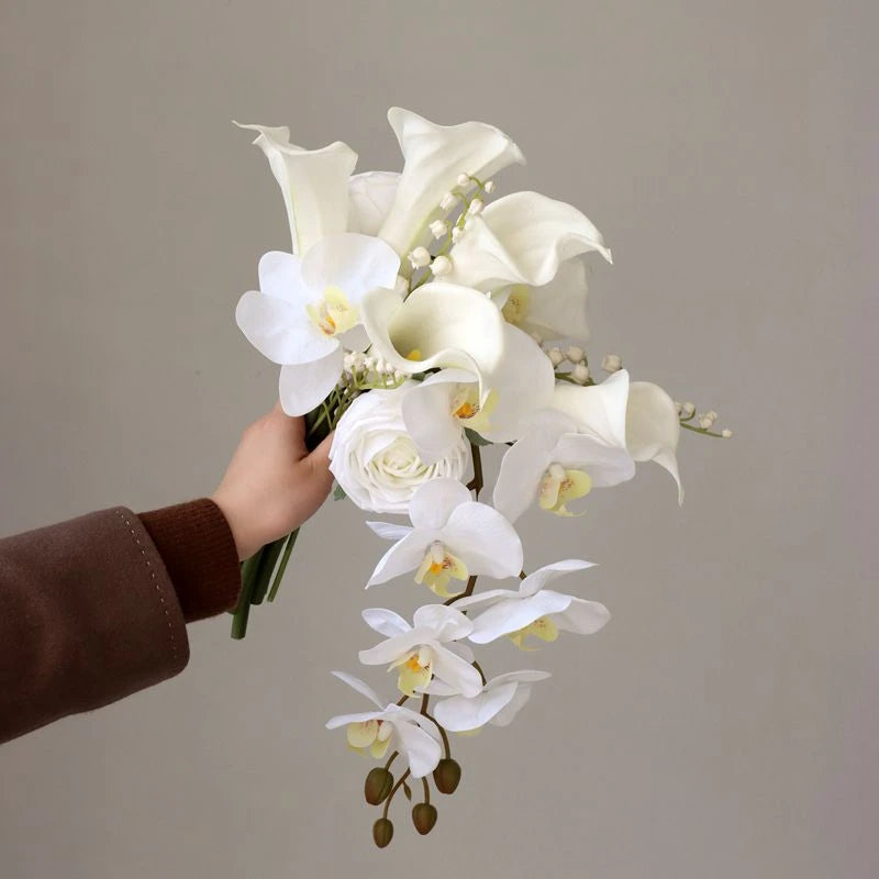 A hand holds a bouquet of elegant white flowers including calla lilies, orchids, and roses against a plain background. The flowers are arranged in a cascading style.