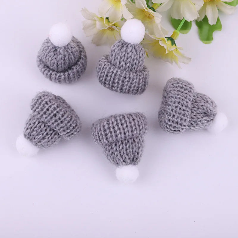 Five small gray knitted hats with white pom-poms arranged on a white surface. Yellow and white flowers are seen in the background.