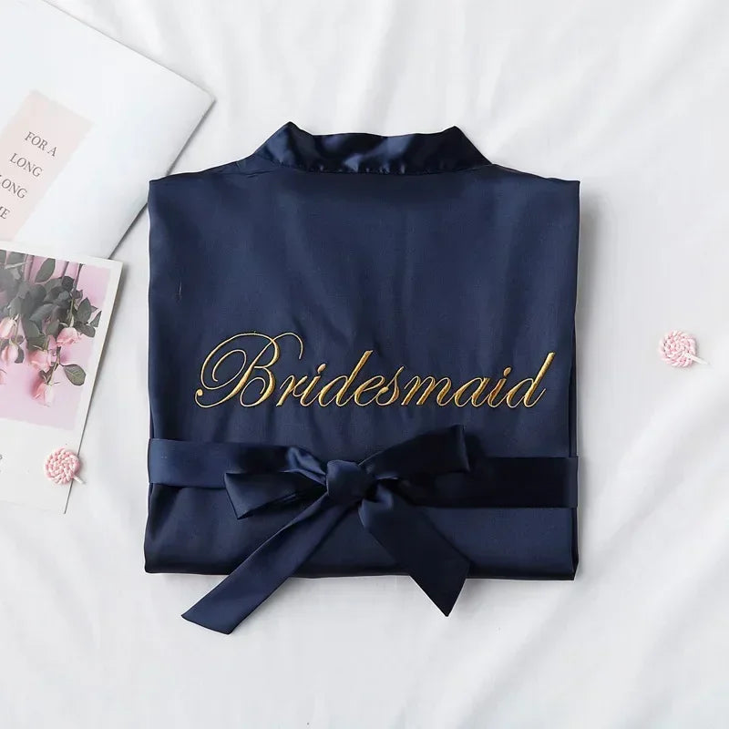 Navy blue bridesmaid robe with gold embroidery and satin bow, lying on a white surface. Nearby, a book with flowers. Two small pink flowers are placed beside it.