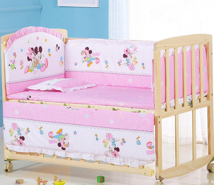 Wooden baby crib with pink bedding featuring cartoon characters. The crib has padded bumpers, a pillow, and is set against a blue wall with toys scattered on the floor.