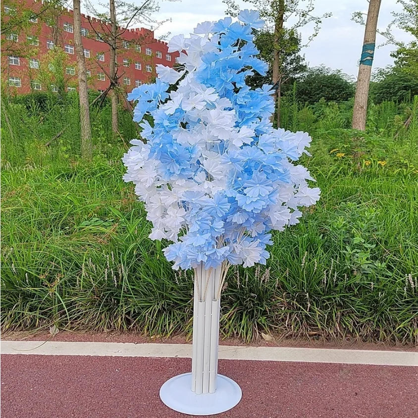 A decorative artificial tree with blue and white flowers stands on a red pathway. It is surrounded by green grass, with a red building and trees in the background.