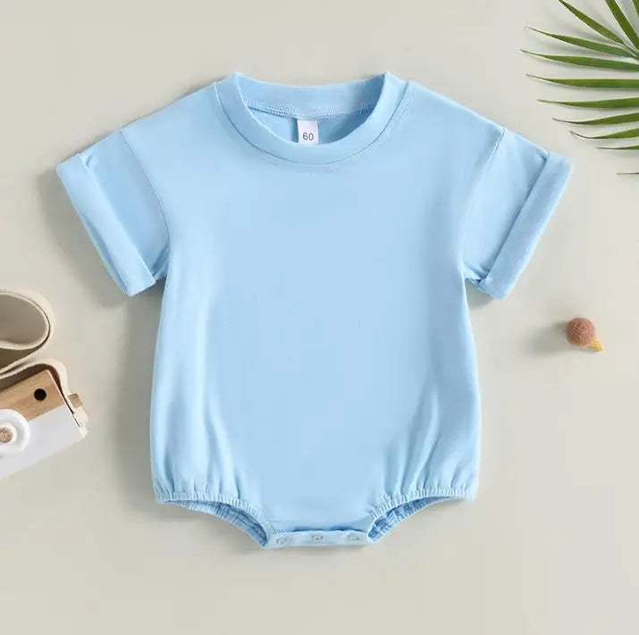 Light blue baby onesie with short sleeves and snap buttons at the bottom, laid flat on a beige surface. There's a small toy camera and a green leaf nearby.
