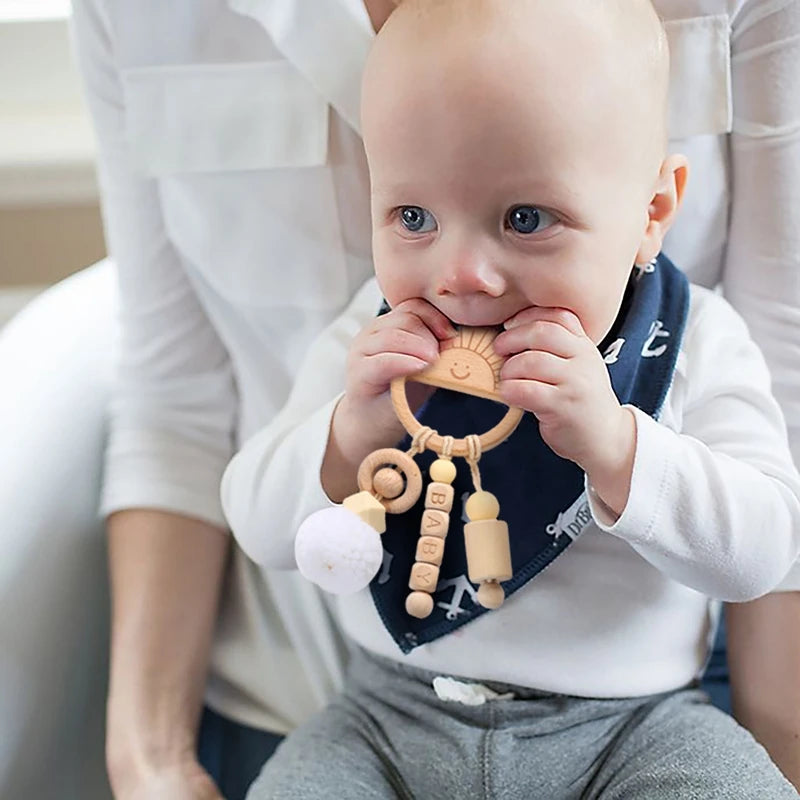 A baby with blue eyes, wearing a navy bib, is seated on an adult's lap, chewing a wooden sun-shaped teething toy with various hanging beads and shapes.