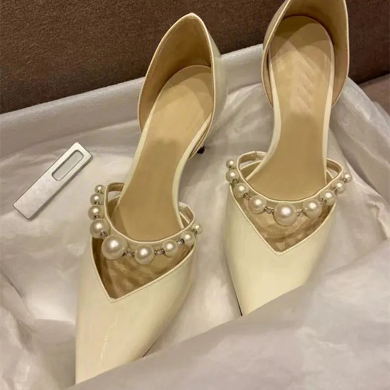 Elegant cream-colored high heels with pointed toes, adorned with pearl and bead straps. The shoes are placed in a box lined with tissue paper.