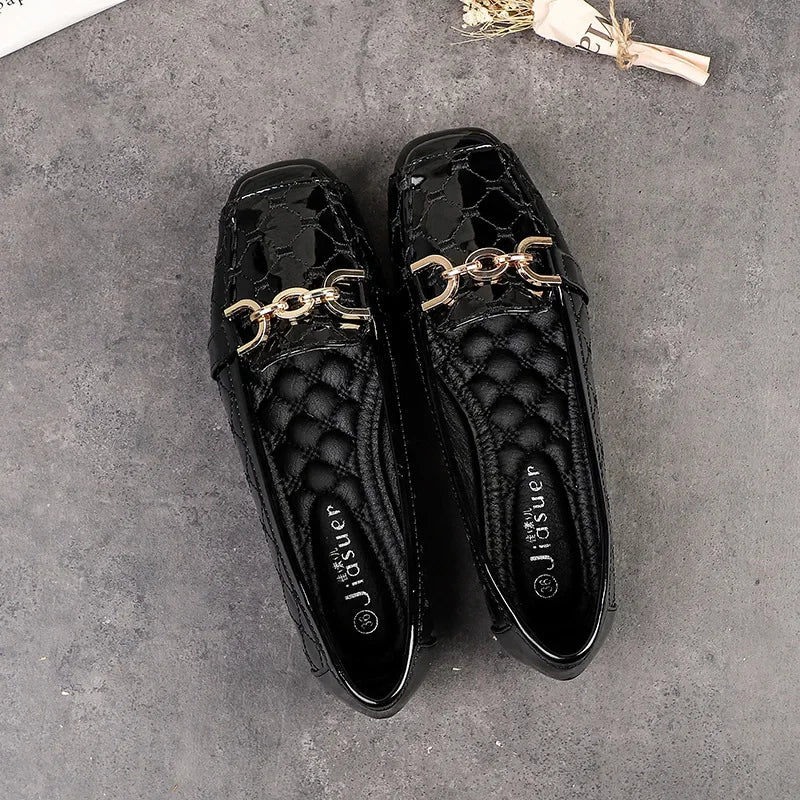 Black patent leather loafers with a crocodile pattern and gold chain detail on the top, placed on a gray surface. A small dried flower bundle is nearby for decoration.