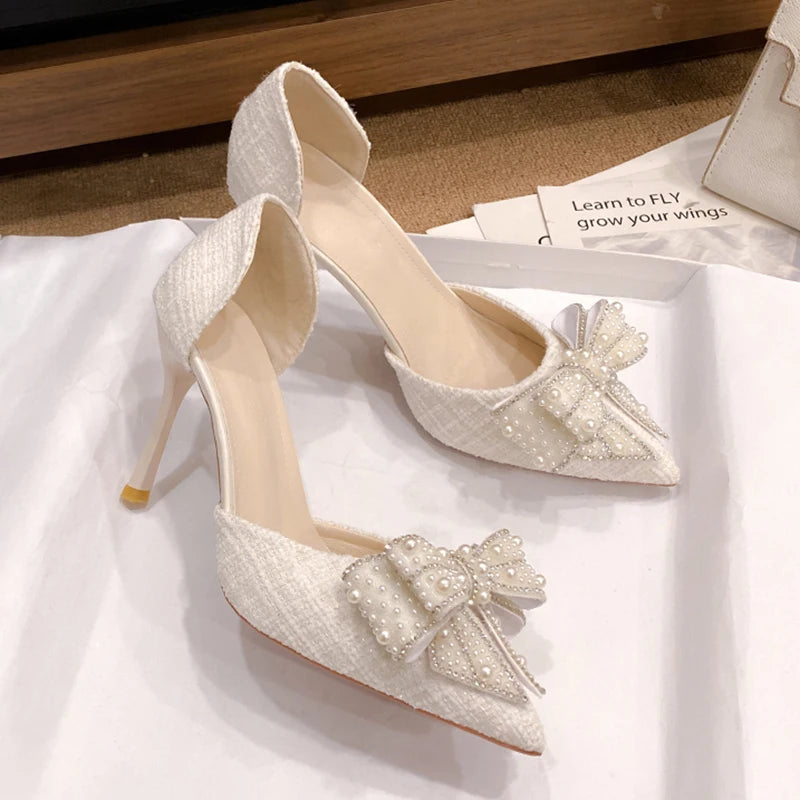 Elegant white high heels with pointed toes, featuring a pearl-embellished bow on each shoe. They rest on a white surface with a book and a handbag in the background.