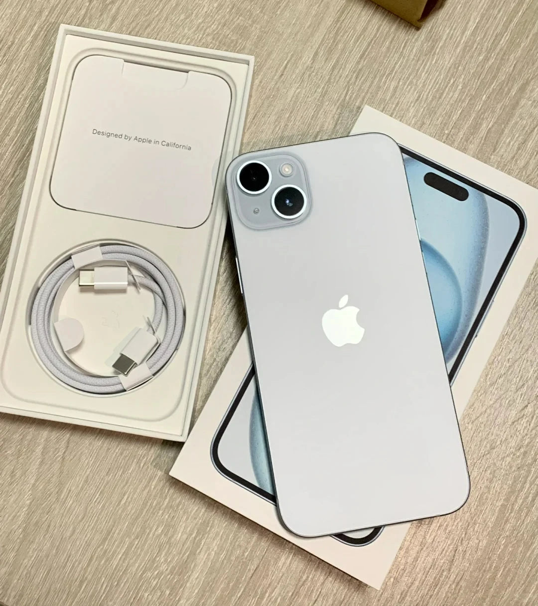 A silver smartphone with dual cameras and Apple logo on the back is atop its box, beside an open package containing a charging cable on a wooden surface.