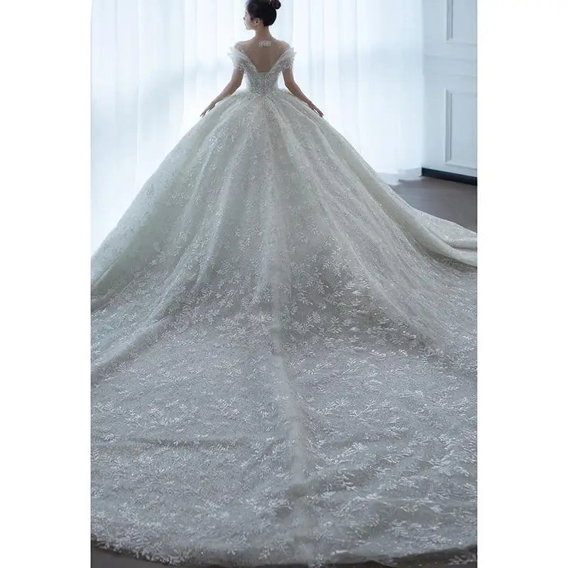 A bride stands in a room wearing an elegant lace wedding gown with a long, flowing train. The dress features intricate floral patterns and an off-shoulder design.