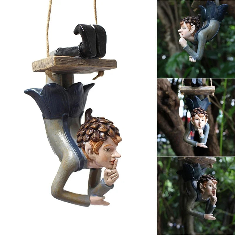 Hanging garden ornament of an elf-like figure with pinecone hat, positioned upside down holding a finger to its lips, attached to a wooden platform. Trees form a green background.