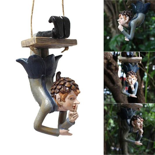 Hanging garden ornament of an elf-like figure with pinecone hat, positioned upside down holding a finger to its lips, attached to a wooden platform. Trees form a green background.