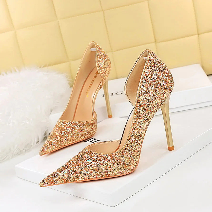 Sparkling gold high heels with pointy toes and stiletto heels on a white surface, against a light cream quilted background, creating a glamorous and elegant look.
