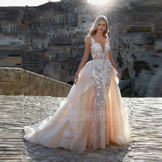 A woman in a flowing, lace-embellished gown stands on a cobblestone path, with a sunlit stone village in the background. The dress features sheer panels and intricate floral details.