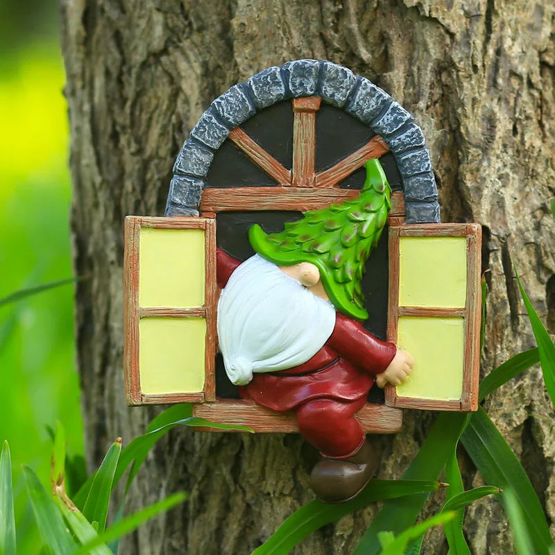 Whimsical garden decoration of a gnome with a green hat climbing through a small window on a tree trunk, surrounded by green foliage.