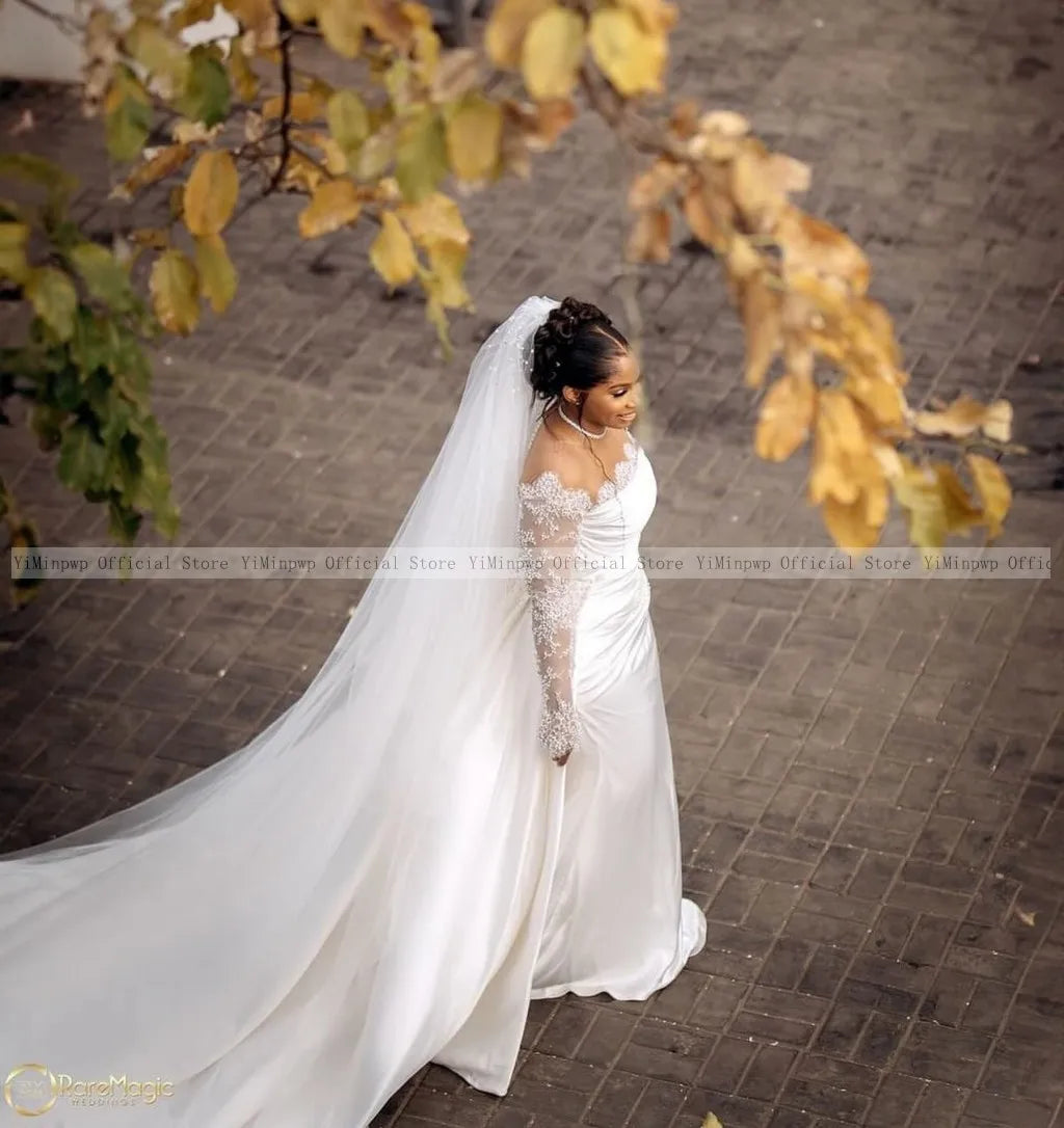 A bride in a satin wedding gown with lace sleeves walks on a paved path, surrounded by autumn leaves. Her long veil flows behind, and her hair is styled in an elegant updo.