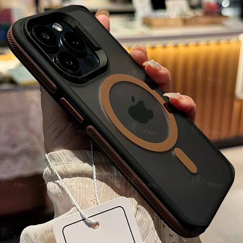 Hand holding a black iPhone with a protective case featuring a brown circle design and a visible logo. Background includes a blurred indoor setting with warm lighting.
