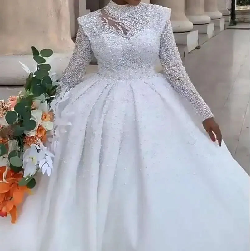 Bride in a sparkling, long-sleeved white gown holds a bouquet of vibrant orange, white, and green flowers. She stands beside stone columns on a paved walkway.