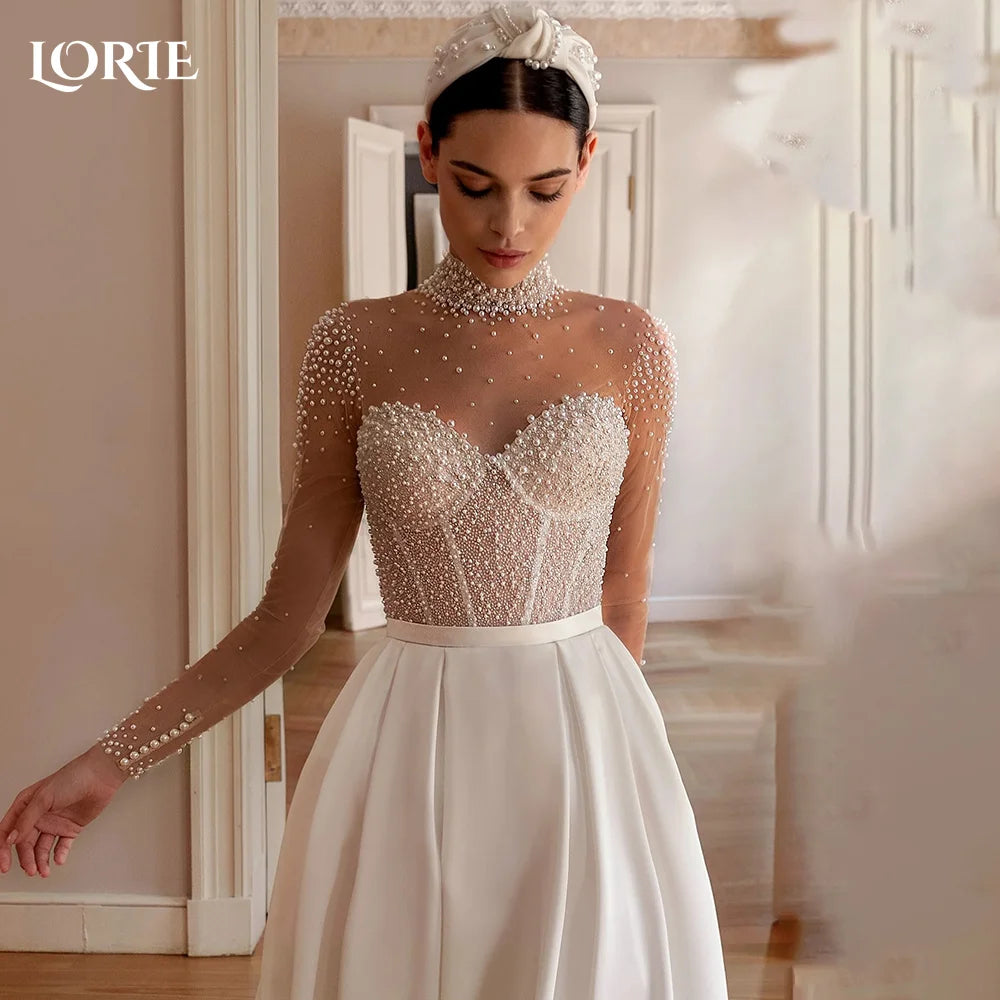 Bride in an elegant wedding dress with a sheer, pearl-embellished bodice and long sleeves. She wears a white headband, standing in a softly lit room with open doors.