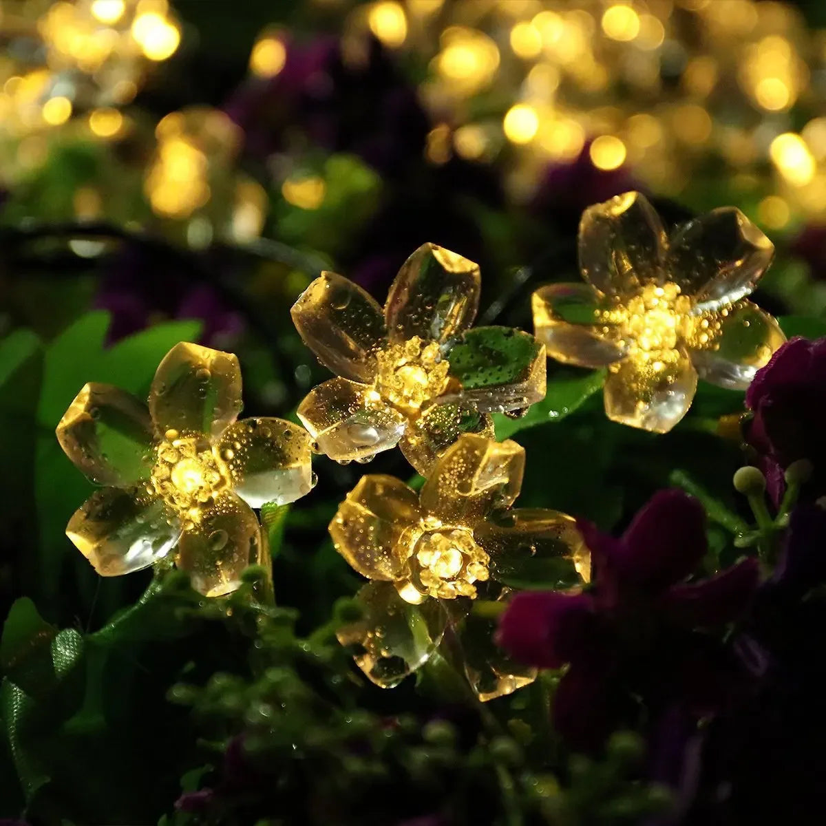 Close-up of illuminated artificial flowers with glowing yellow lights, resembling real blossoms. They are surrounded by green foliage, creating a warm and vibrant display.